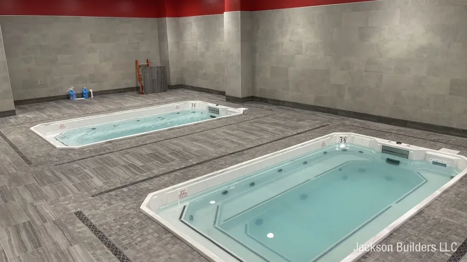 Two HydroWorx aquatic therapy pools installed side by side in the Jacksonville State University athletic training room, set flush into grey porcelain tile with linear trench drains and the school's red accent band along the upper wall.